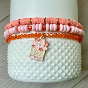 Puede incluir: Tres pulseras superpuestas con cuentas rosas y naranjas y un dije dorado que dice "Plant Mama" con una flor rosa en la parte superior.