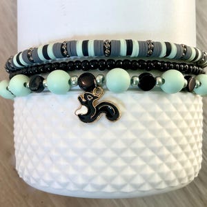 May include: A collection of beaded bracelets in shades of black, grey, and mint green. One bracelet has a skunk charm. The bracelets are presented on a white, textured surface.