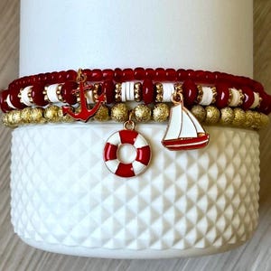 May include: Stacked nautical-themed bracelets with red, white, and gold beads. Charms include a red anchor, a life preserver, and a sailboat. The bracelets are displayed on a white textured surface.