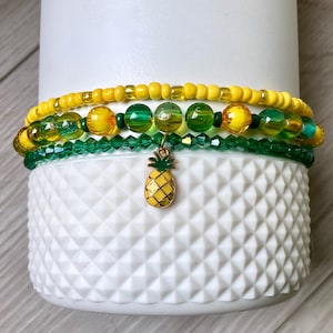 Könnte beinhalten: Ein Set aus drei Armbändern mit gelben und grünen Perlen, inklusive einem Ananas-Anhänger. Die Armbänder sind auf einer weißen Keramiktasse mit strukturierter Oberfläche gestapelt.