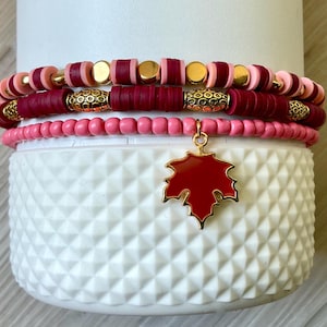 May include: A set of stacked bracelets in pink, burgundy, and gold. The top bracelet has pink and gold beads, the middle one has burgundy and gold beads, and the bottom one is pink. A red leaf charm hangs from the bottom bracelet.