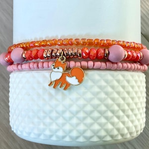 May include: Three beaded bracelets in shades of orange, pink, and rose gold. The top bracelet is orange, the middle is pink with rose gold accents, and the bottom is light pink with a fox charm. The bracelets are stacked on a white textured surface.