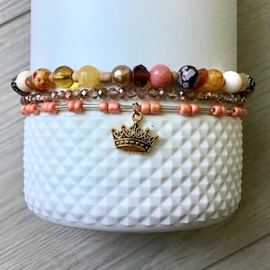 Könnte beinhalten: Ein Set aus vier Armbändern mit einem goldenen Kronenanhänger. Die Armbänder bestehen aus verschiedenen Perlen, darunter rosa, orange, gelb und braun. Die Armbänder sind übereinander gestapelt.