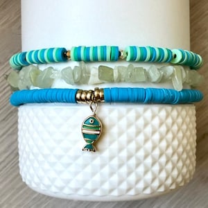 Könnte beinhalten: Drei Armbänder auf einer weißen Oberfläche. Das oberste Armband hat grün-blaue Streifenperlen. Das mittlere Armband hat hellgrüne Steinperlen. Das untere Armband ist blau mit einem goldenen Akzent und einem Fischanhänger.