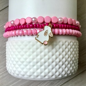 Könnte beinhalten: Ein Set aus vier rosafarbenen und weißen Perlenarmbändern mit einem weißen Hasenanhänger. Die Armbänder sind auf einer weißen, strukturierten Oberfläche gestapelt.