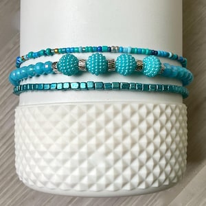 Op de afbeelding: Vier turquoise blauwe kralenarmbanden gestapeld op elkaar. De armbanden zijn gemaakt van kleine kralen en grotere ronde kralen. De armbanden zijn op een witte achtergrond.