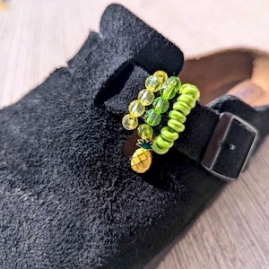Könnte beinhalten: Ein schwarzer Wildleder-Clog mit einem dekorativen Riemen-Accessoire. Das Accessoire besteht aus zwei Strängen aus durchsichtigen gelben und grünen Perlen, einem lindgrünen spiralförmigen Band und einem kleinen gelb-grünen Ananas-Anhänger.