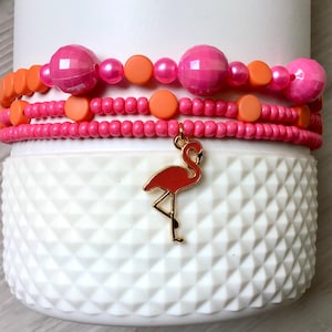 May include: A stack of beaded bracelets in shades of pink and orange, with a gold-colored flamingo charm. The bracelets are on a white, textured surface. The bracelets feature various bead sizes and shapes.