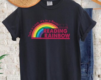May include: Black t-shirt with a rainbow graphic and the text "Take a Look, it's in a Book! READING RAINBOW". The design features a rainbow with red, orange, yellow, green, blue, and purple colors. The shirt is hanging on a wooden hanger.