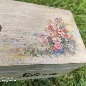 Può includere: Una scatola di legno con un disegno floreale dipinto sulla parte superiore. La scatola è dipinta in un colore beige chiaro con una finitura usurata. Il disegno floreale è in tonalità di rosa, rosso e giallo. La scatola ha il nome "Silvia" scritto sul davanti.