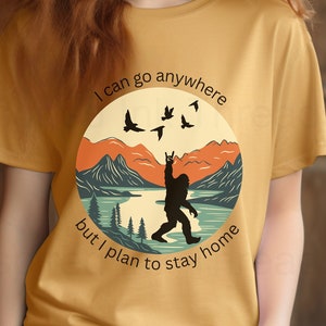 May include: A yellow t-shirt with a retro-style graphic of a Bigfoot standing in a mountain landscape with the text "I can go anywhere but I plan to stay home".