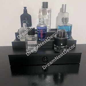 May include: A black tiered shelf display with six glass bottles of cologne. The bottles are arranged in two rows. The top row has three bottles, and the bottom row has three bottles. The bottles are all different sizes and shapes. The shelf is made of wood and has a black finish.