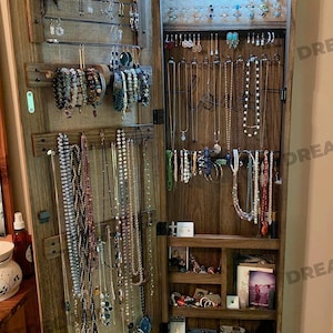 Può includere: Un'armadio portagioie in legno con la porta aperta, che mostra varie collane, bracciali e orecchini. L'interno presenta più ganci e ripiani per riporre gli oggetti. L'armadio è in legno scuro e ha un'estetica rustica.