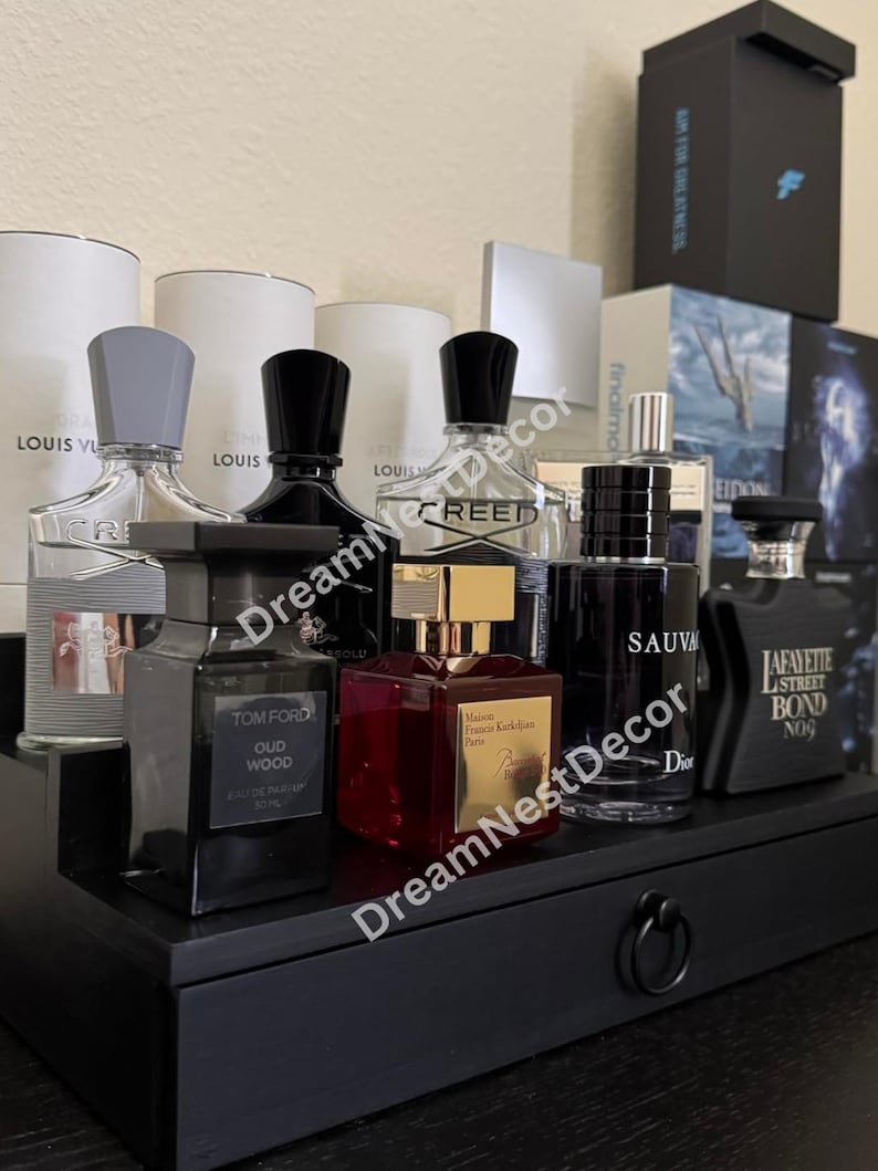 May include: A black wooden shelf with a drawer and a ring pull handle, displaying a collection of various designer perfume bottles. The bottles are arranged in a row and include brands such as Creed, Tom Ford, and Maison Francis Kurkdjian.