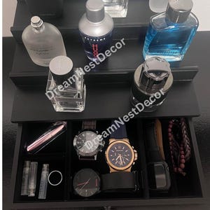 May include: A black tiered organizer with a drawer. The top tier has five perfume bottles, including a clear bottle with the word "FIERCE" on it. The drawer has a black velvet lining and contains a variety of items, including watches, a bracelet, and a small pink lipstick.
