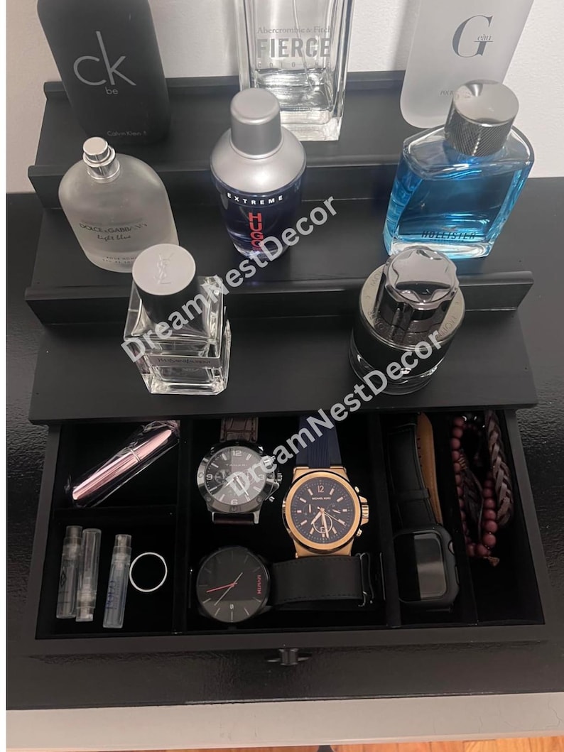 May include: A black tiered organizer with a drawer. The top tier has five perfume bottles, including a clear bottle with the word "FIERCE" on it. The drawer has a black velvet lining and contains a variety of items, including watches, a bracelet, and a small pink lipstick.