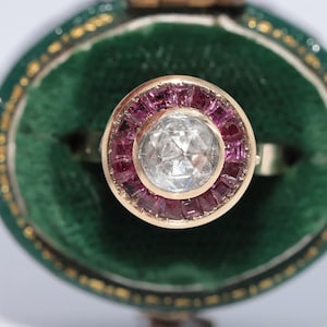 May include: A gold ring featuring a central clear gemstone encircled by a halo of square, pink gemstones. The ring is displayed against a green velvet backdrop within a jewellery box.