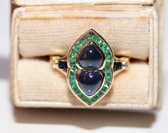 Può includere: Un anello d'oro dal design unico, con due pietre preziose blu scuro a forma di cuore, circondate da un bordo di piccole pietre verdi. L'anello è presentato in un portagioie in stile vintage.