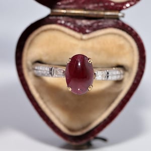 Op de afbeelding: Een ring met een centrale ovale cabochon geslepen dieprode edelsteen in gouden zettingen, gemonteerd op een zilverkleurige band met kleine rechthoekige heldere stenen aan de zijkanten, gepresenteerd in een hartvormig donkerrood juwelendoosje.
