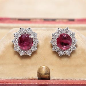 Puede incluir: Un par de pendientes de botón con rubíes rojos de talla ovalada en el centro, cada uno rodeado por un halo de pequeños diamantes blancos redondos y transparentes engastados en un marco metálico. Los pendientes se presentan en un joyero rojo.