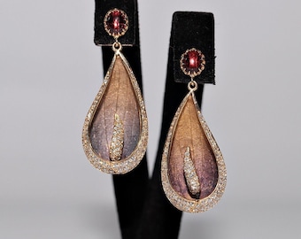 May include: A pair of gold drop earrings with a teardrop-shaped design. The earrings feature a brown and purple gradient with a clear crystal in the center. The earrings are accented with a red gemstone at the top.