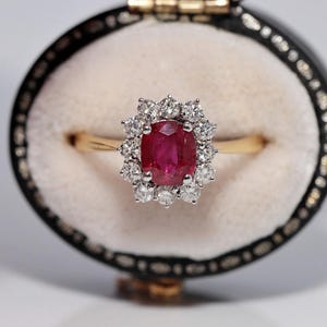 Op de afbeelding: Een gouden ring met een centrale ovaal geslepen rode robijn, omringd door een halo van twaalf rond geslepen witte diamanten. De ring wordt gepresenteerd in een zwart, ovaalvormig juwelendoosje met een crèmekleurige fluwelen voering.