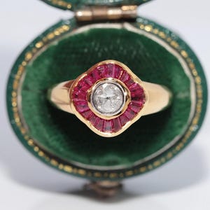 Puede incluir: Un anillo de oro con un diamante blanco redondo central rodeado por un halo de rubíes rojos de talla baguette dispuestos en forma de flor festoneada. El anillo se presenta dentro de un joyero ovalado de color verde oscuro con forro de terciopelo.