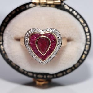 May include: A heart-shaped ring featuring a central pear-cut ruby surrounded by a halo of channel-set baguette rubies and an outer border of small round diamonds, all set in a gold band, presented within a round black velvet-lined jewelry box.