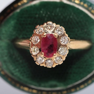 May include: A gold ring featuring a central oval-cut red ruby surrounded by a halo of ten round-cut white diamonds. The gemstone setting is mounted on a polished gold band, creating a classic cluster design with a vibrant red and white contrast.
