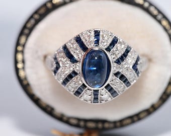 May include: A silver ring with a large oval blue gemstone at the center, surrounded by smaller blue and clear stones. The ring has a geometric design with a vintage aesthetic, set against a cream-colored background.