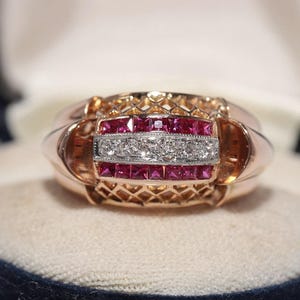 May include: A gold ring with a rectangular center section. The center features a row of small diamonds between two rows of square red gemstones. The ring is set in a blue velvet jewelry box.