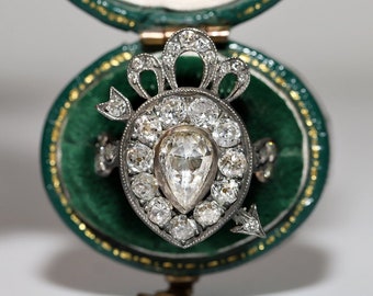 May include: A pear-shaped diamond ring set in silver with a decorative bow and arrow design. The ring is surrounded by smaller diamonds and is in a green velvet ring box.