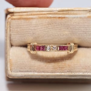 Pode incluir: Um anel de ouro com uma fila horizontal de quatro rubis vermelhos de corte quadrado e dois diamantes transparentes de corte redondo no centro. As pedras estão cravadas num canal de ouro polido com barras decorativas nas extremidades.