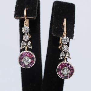May include: A pair of drop earrings featuring a circular design with a central round diamond surrounded by a halo of square-cut magenta rubies. Above the circular drop are two smaller round diamonds and a leaf-shaped metal accent in gold.