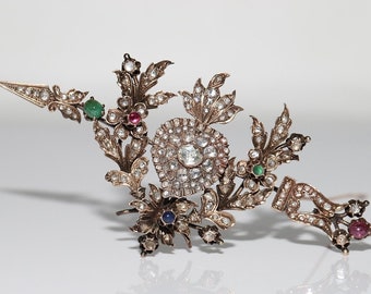 May include: An antique gold-coloured hair comb with a floral design. The comb features numerous clear gemstones and small coloured gemstones, including emeralds, rubies, and sapphires. The design includes a central cluster of gemstones and delicate leaf and flower details.