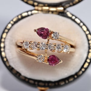 Op de afbeelding: Een gouden ring met een uniek ontwerp, met twee peervormige roze edelstenen en een rij kleine, ronde, heldere edelstenen. De ring wordt gepresenteerd in een zwart-gouden sieradendoos met een crèmekleurig interieur.