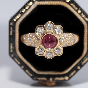 Op de afbeelding: Een gouden ring met een bloemvormig ontwerp. Het middenstuk heeft een ronde, dieprode edelsteen, omringd door witte diamanten. De band is ook versierd met kleinere diamanten, gepresenteerd in een zwarte fluwelen sieradendoos.