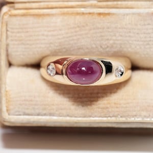 Può includere: Un anello d'oro con una grande gemma di rubino ovale al centro, affiancata da due diamanti più piccoli. L'anello è incastonato in una fascia d'oro lucida e viene presentato in un portagioie di velluto beige.