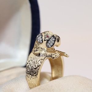 Può includere: Anello color oro con un design dettagliato di un carlino. Gli occhi del cane sono accentuati da piccole pietre bianche e una singola pietra rossa è incastonata al centro della fronte. L'anello è presentato in un cofanetto di velluto blu.