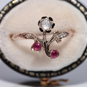 Puede incluir: Un anillo antiguo con diseño floral. El anillo presenta un diamante central, dos acentos de rubí y diamantes más pequeños engastados en una banda de oro. El anillo se exhibe en un joyero negro y dorado con interior color crema.