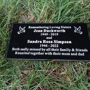 May include: Black granite memorial plaque with white text remembering two sisters, Jean Duckworth (1949-2019) and Sandra Rosa Simpson (1946-2022). The plaque includes the text "Remembering Loving Sisters" and "Both sadly missed by all their family & friends. Reunited together with their mum and dad."