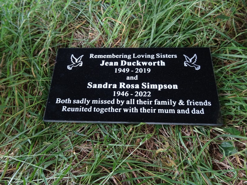 May include: Black granite memorial plaque with white text remembering two sisters, Jean Duckworth (1949-2019) and Sandra Rosa Simpson (1946-2022). The plaque includes the text "Remembering Loving Sisters" and "Both sadly missed by all their family & friends. Reunited together with their mum and dad."