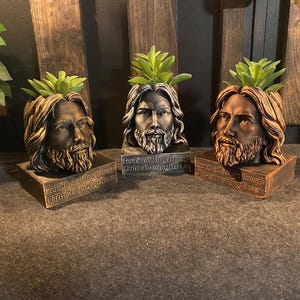 Könnte beinhalten: Drei bronzefarbene Jesus-Kopf-Pflanzgefäße, jeweils mit einer grünen Sukkulente. Die Pflanzgefäße stehen auf rechteckigen Sockeln mit dem Text "I can do all things through Christ who strengthens me."