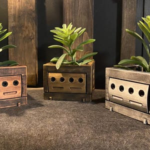 May include: Three wooden planter boxes with different finishes. The boxes are shaped like the Nintendo GameCube console and have slots for game cartridges. Each box has a small plant growing inside.