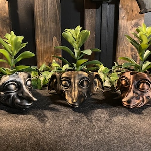 May include: Three ceramic houseplant pots shaped like the head of a goblin or elf with large ears. The pots are painted in black, brown, and bronze. Each pot has a small green succulent plant growing inside.
