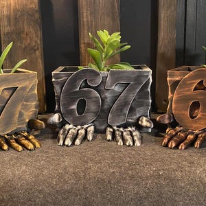 Op de afbeelding: Drie decoratieve plantenbakken met het nummer "67". De plantenbakken worden ondersteund door handvormige bases en bevatten kleine groene planten. De plantenbakken zijn in brons, grijs en bruin. Ideaal voor een tuin.