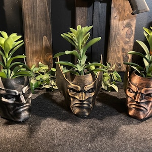 May include: Three ceramic planters shaped like Wolverine's head, each with a different paint finish: black, gold, and bronze. Each planter is filled with a small green succulent plant.