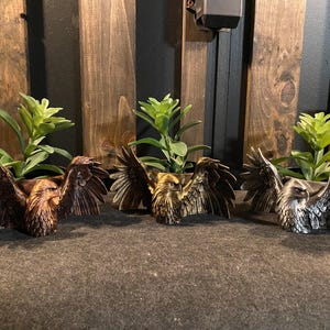 May include: Three decorative planters with eagle designs. Each planter features a detailed eagle with outstretched wings, holding a small green plant. The planters are in bronze, gold, and silver tones, creating a striking display.