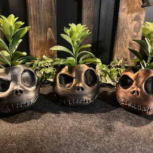 以下が含まれることがあります： 緑色の多肉植物が入った、黒と金のジャック・スケリントン柄のセラミック製プランター3つ。プランターは、使い古したような仕上げになっています。