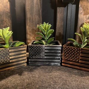 May include: Three wooden planter boxes with an American flag design. Each planter box has a different color scheme: black, brown, and copper. The boxes are filled with green succulents.